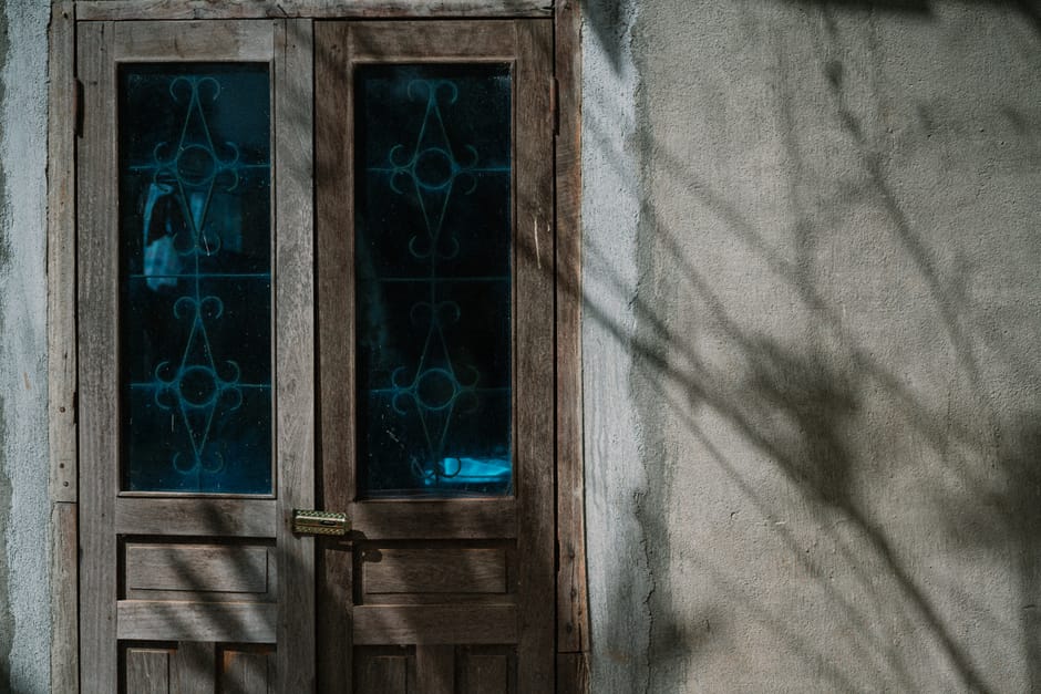 A rustic wooden door with artistic glass panels, sunlit with shadows cast on adjacent wall.
