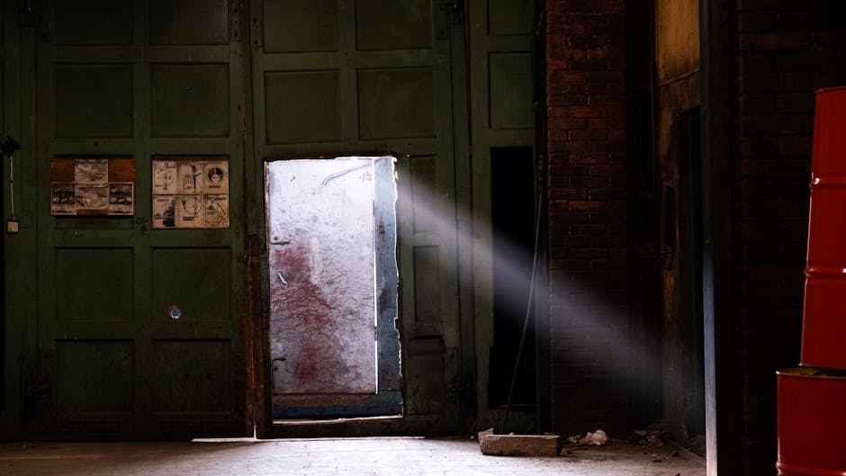A beam of light shines through a partially open door in an abandoned industrial space.