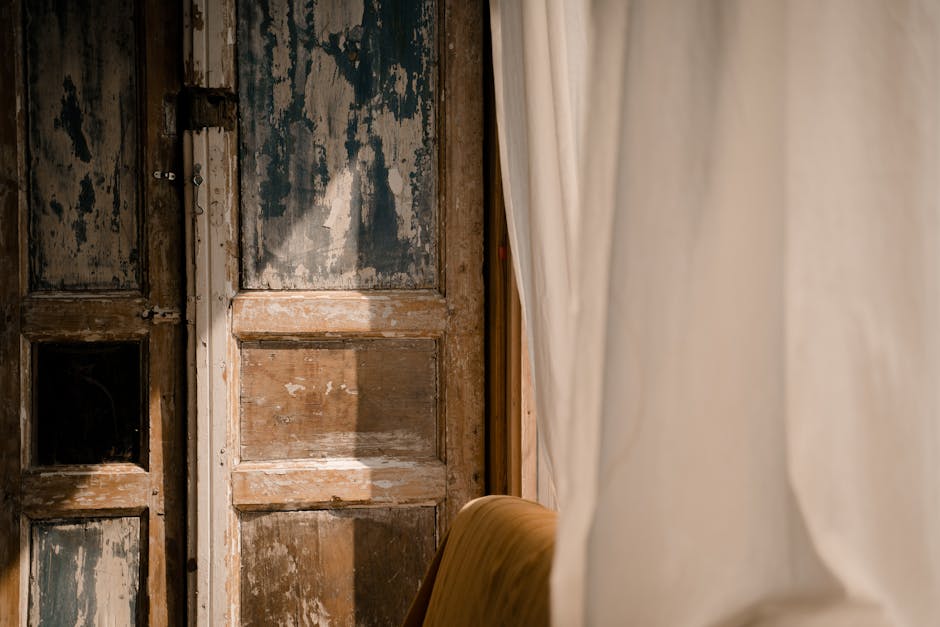 Sunlit rustic wooden door with aged texture partially covered by flowing curtains.